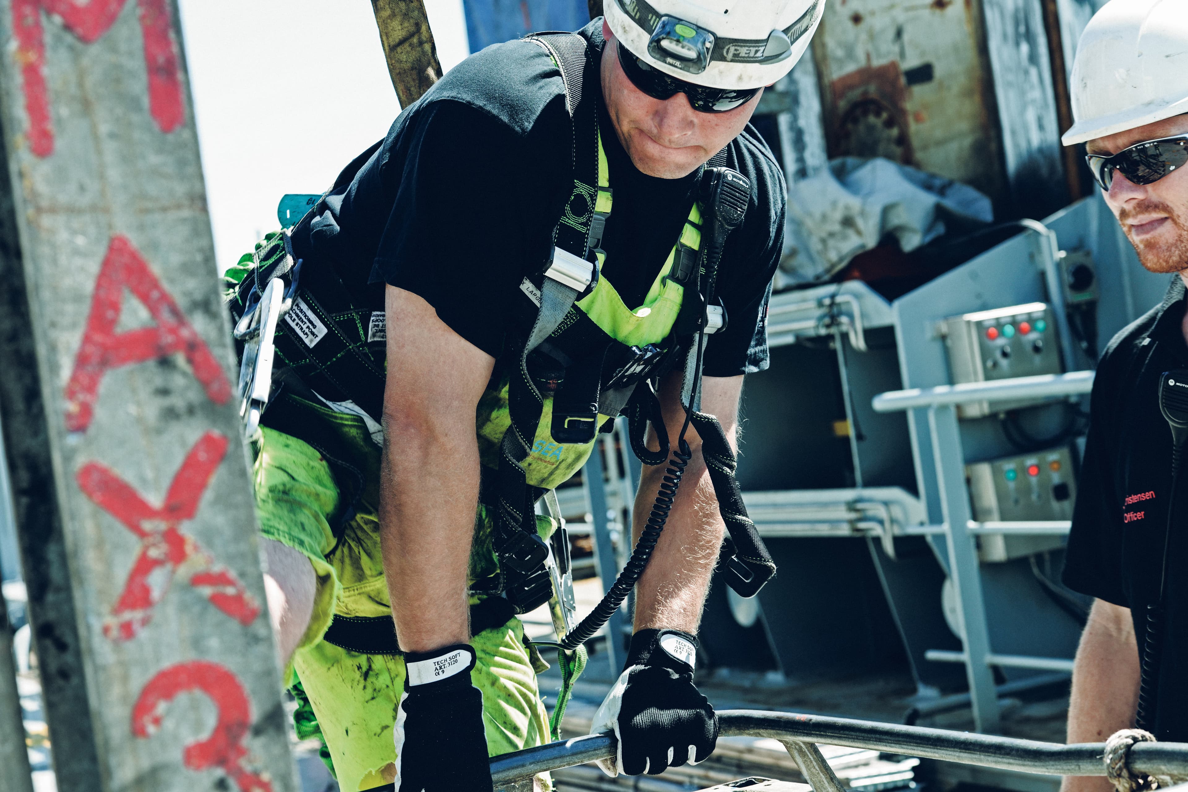A worker wearing protective gear and a harness, engaged in a hands-on task, highlighting safety practices and teamwork in a high-risk industrial environment.