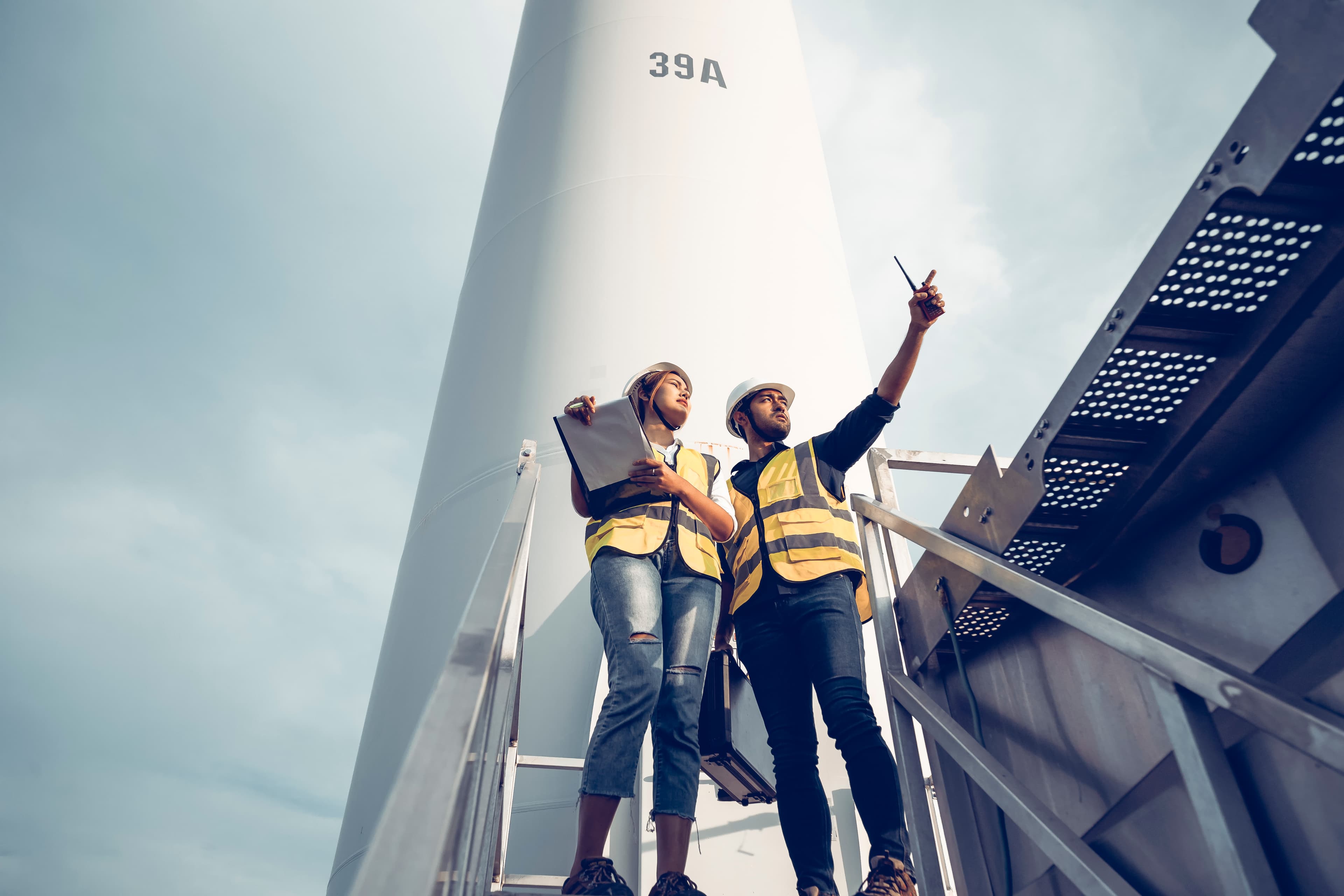 Two engineers in high-visibility vests conduct an onsite inspection at the base of a wind turbine, reviewing details and pointing out observations.