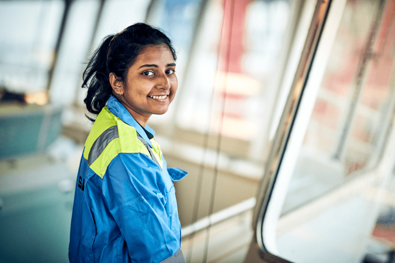 a woman in a blue and yellow work uniform smiling in what appears to be a ship's bridge or control room. The setting suggests she is a seafarer or maritime professional, reflecting a confident and positive attitude in her work environment