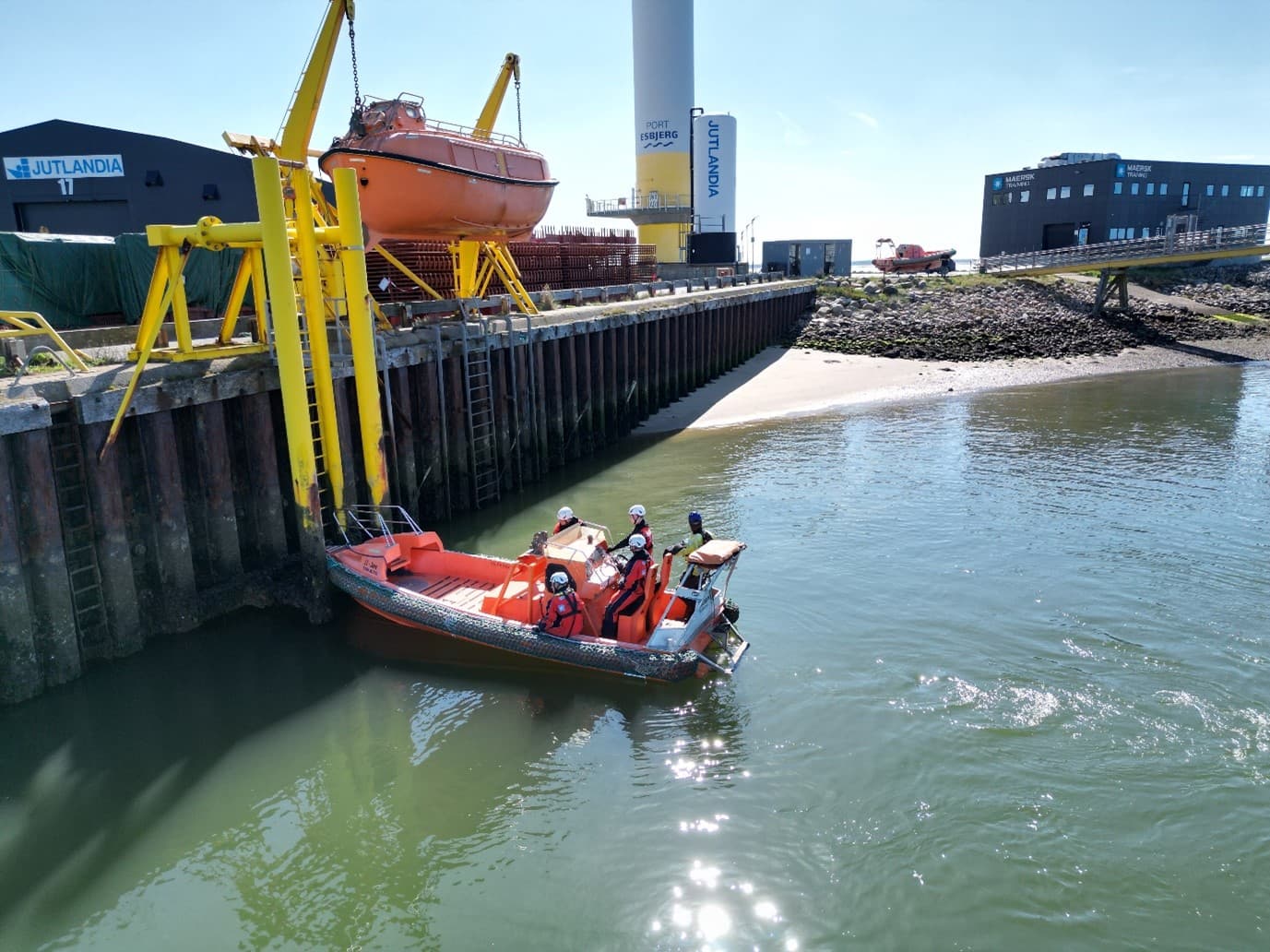 The image shows a small boat with three people in life jackets near a dock, with a yellow crane holding an orange lifeboat. The setting appears to be an industrial port, likely for safety at sea training.