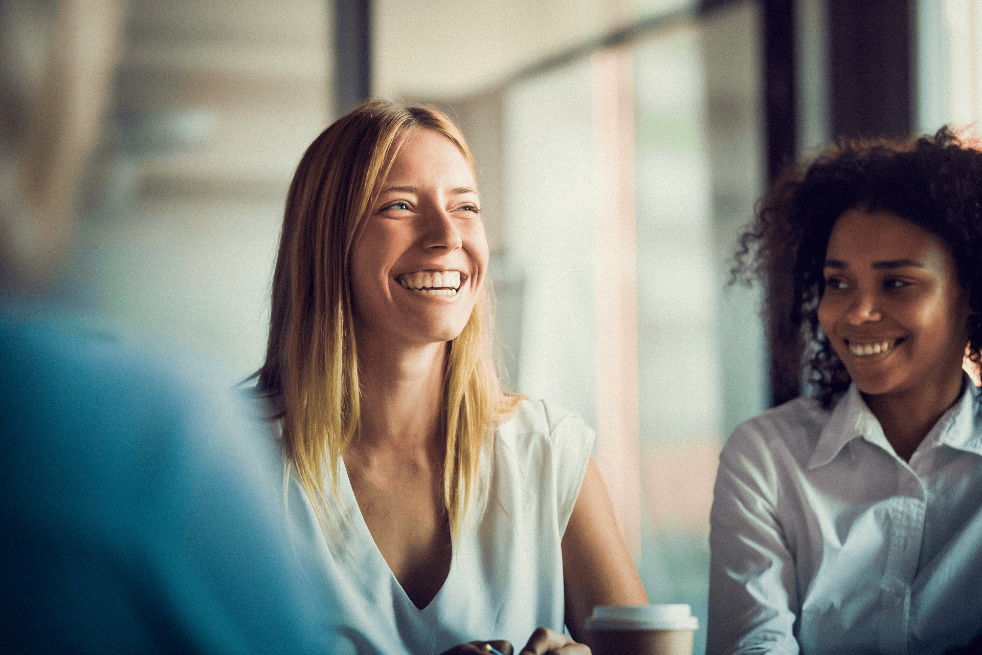 The image shows two women smiling and engaging in conversation, seated in a bright, modern office setting